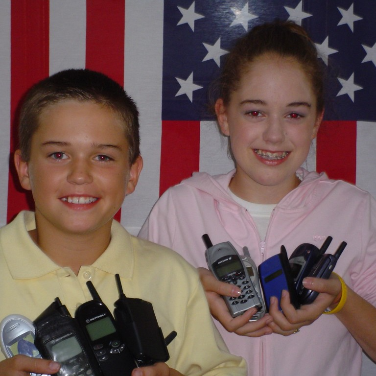 Young Brittany and Robbie Bergquist holding multiple cell phones, smiling in front of an American flag.