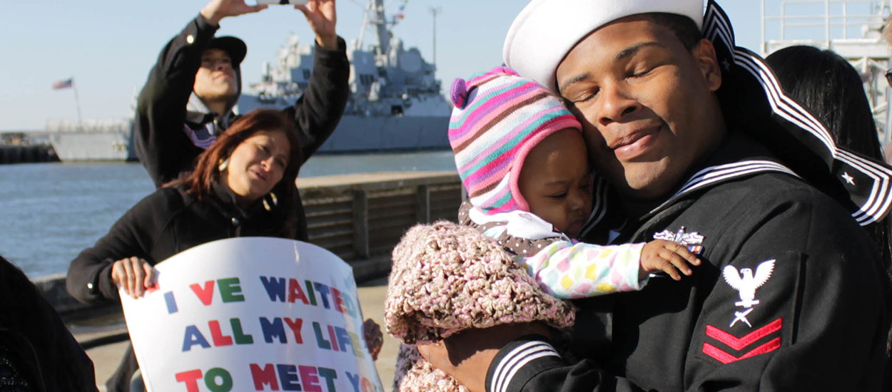 A U.S. Navy sailor embraces his child upon returning home, with a family member holding a sign reading "I've waited all my life to meet you".
